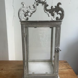 May include: A decorative metal lantern with clear glass panels. The lantern has an ornate, scrollwork design at the top and a small handle. The frame is a distressed grey colour, and the lantern sits on a wooden surface.