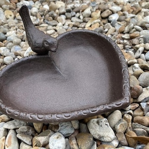 May include: A heart-shaped cast iron bird bath with a bird figurine on top. The bath is brown and has a decorative edge.