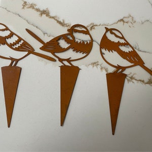 May include: Three rusty metal bird silhouettes on stakes. The birds are facing different directions and have a simple, cut-out design.