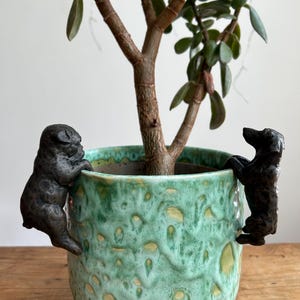 May include: Two black ceramic dog figurines climbing the side of a green ceramic planter with a leafy green plant growing inside.