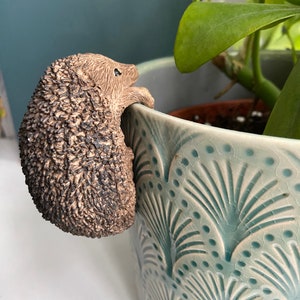 K&ouml;nnte beinhalten: Eine braune Keramik-Igelfigur, die an der Seite eines blau-wei&szlig; gemusterten Blumentopfs hochklettert.