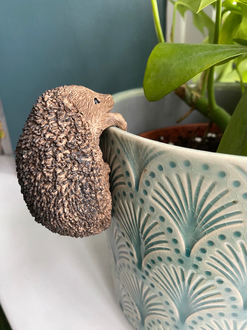 K&ouml;nnte beinhalten: Eine braune Keramik-Igelfigur, die an der Seite eines blau-wei&szlig; gemusterten Blumentopfs hochklettert.
