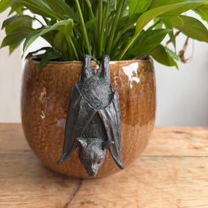 May include: A dark grey, textured bat figurine hanging upside down from the rim of a round, glossy, amber-coloured ceramic planter. The bat has detailed wings folded against its body and pointed ears, while the planter contains a green plant.