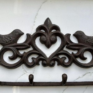 May include: A cast iron wall mounted key holder with a decorative design featuring two birds and a heart. The holder has three hooks for hanging keys.