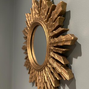 K&ouml;nnte beinhalten: Ein goldener Sonnenspiegel mit einer runden, reflektierenden Oberfl&auml;che. Der Spiegel hat einen dekorativen, strukturierten Rahmen mit einem Sonnenstrahlendesign.