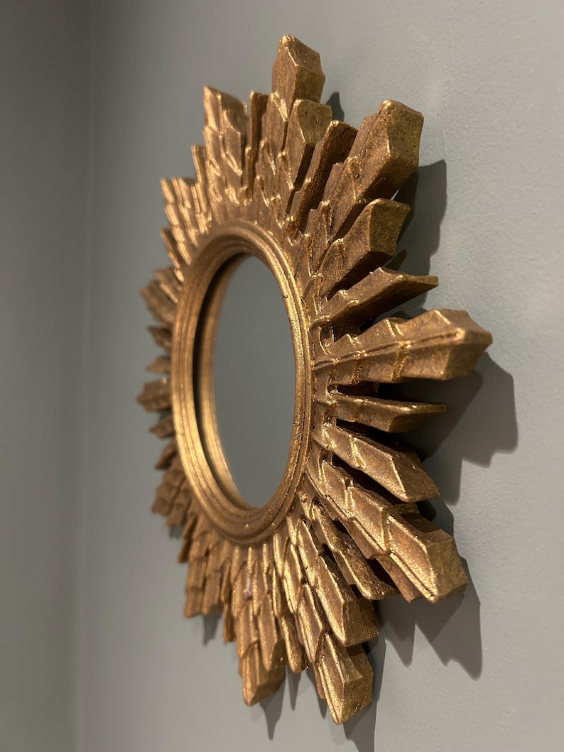 K&ouml;nnte beinhalten: Ein goldener Sonnenspiegel mit einer runden, reflektierenden Oberfl&auml;che. Der Spiegel hat einen dekorativen, strukturierten Rahmen mit einem Sonnenstrahlendesign.