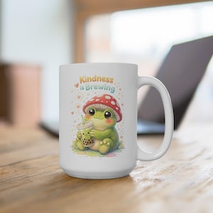 May include: White ceramic mug featuring a cartoon frog wearing a mushroom cap and holding a boba tea. The mug has the text "Kindness is Brewing" with hearts. The mug is on a wooden surface.