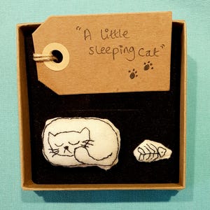 May include: A square cardboard gift box with a black velvet insert containing a white fabric patch embroidered with a sleeping cat and a small fish skeleton. A brown paper tag with the handwritten text A little sleeping cat is attached.