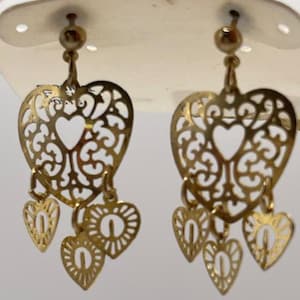 May include: Gold-toned filigree heart earrings. Each earring features a large heart-shaped design with intricate cut-out patterns and a smaller heart pendant. The earrings are suspended from a post.
