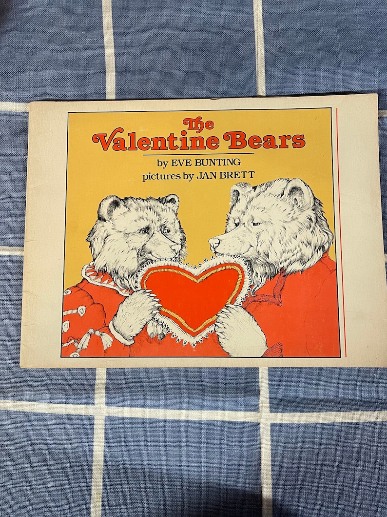 May include: The cover of a children's book titled "The Valentine Bears" by Eve Bunting, with illustrations by Jan Brett. The cover features two bears, one white and one brown, holding a red heart-shaped object.
