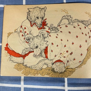 May include: Two white bears wearing red and white patterned dresses are lying on a bed of straw. The bear on the left is sitting up and looking at the bear on the right.