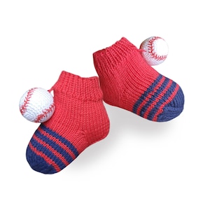 May include: A pair of red knitted baby booties with blue stripes and a white baseball pom-pom on each bootie.