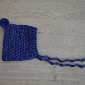 Op de afbeelding: Een blauwe gehaakte baby muts met een pompon bovenop en strikjes.