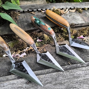 May include: Four handcrafted garden trowels with wooden handles and metal blades. The handles vary in colour and design, with some featuring inlaid decorative elements. The trowels are arranged on a weathered wooden surface.