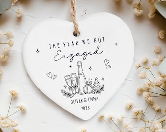 May include: A white heart-shaped ceramic ornament hanging by a jute string. It features black line art of a champagne bottle, two glasses, a diamond ring, and a tree, with the text "THE YEAR WE GOT Engaged", "OLIVER & EMMA", and "2026".