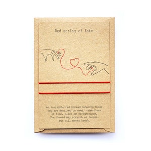 May include: A kraft paper card with the text "Red string of fate" and a drawing of two hands connected by a red string forming a heart. Two red strings are attached to the card. The card also includes the text: "An invisible red thread connects those who are destined to meet..."