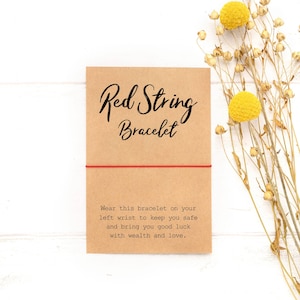 May include: Red string bracelet on a brown card with the text "Red String Bracelet" and "Wear this bracelet on your left wrist to keep you safe and bring you good luck with wealth and love."