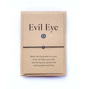 May include: A brown card with the text "Evil Eye" and a blue evil eye charm. A black string bracelet with a blue evil eye charm is attached to the card. The card reads "Wear this bracelet on your wrist to keep you safe and bring you good luck with wealth and love."