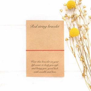 May include: Red string bracelet on a brown card with the text "Red string bracelet" and "Wear this bracelet on your left wrist to keep you safe and bring you good luck with wealth and love."