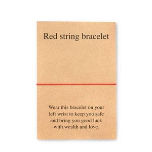 May include: Red string bracelet with a message on a brown paper card. The message reads: "Wear this bracelet on your left wrist to keep you safe and bring you good luck with wealth and love."