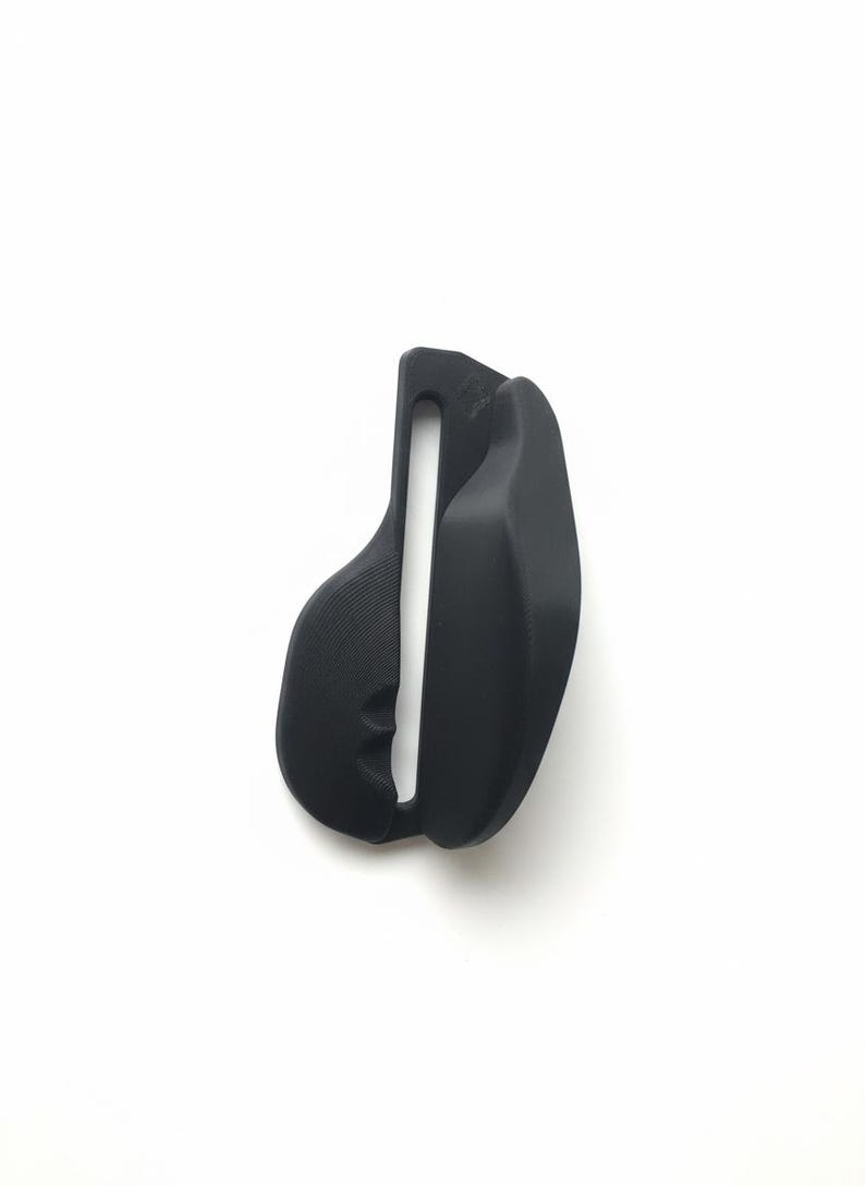 Pode incluir: Um objeto preto em forma de sela com uma abertura vertical central. O objeto tem uma superf&iacute;cie texturizada de um lado e um acabamento liso do outro. O design sugere um foco no conforto e no suporte.
