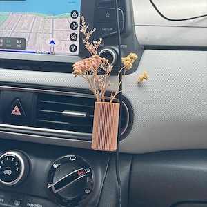 May include: A small, brown, ribbed vase holding dried flowers is clipped to a car's air vent. The car's dashboard is visible, including the navigation screen and climate control dials. The vase adds a touch of natural decor to the vehicle's interior.