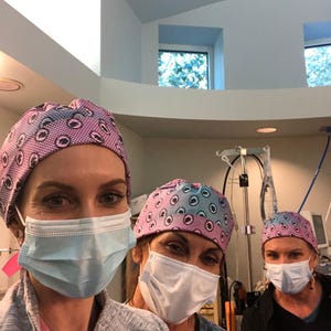 May include: Three people wearing surgical caps and face masks. The caps have a repeating pattern of blue and pink eye-like shapes. The people are in a medical setting, with surgical equipment visible in the background.