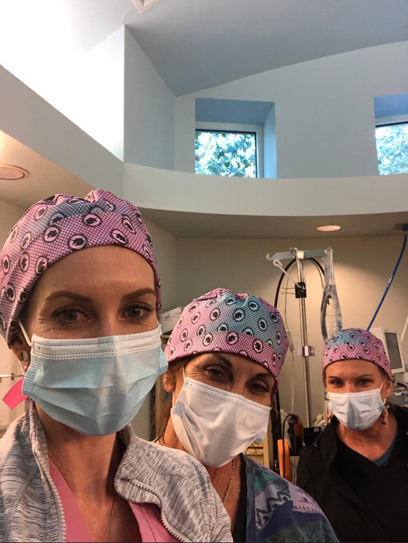 May include: Three people wearing surgical caps and face masks. The caps have a repeating pattern of blue and pink eye-like shapes. The people are in a medical setting, with surgical equipment visible in the background.