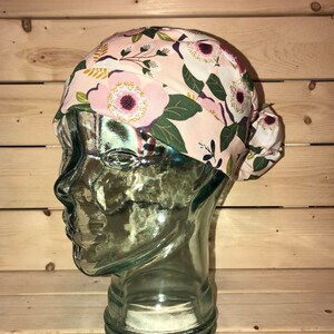 May include: A pink floral print surgical scrub hat with a white and green floral pattern. The hat is tied at the back with a bow.
