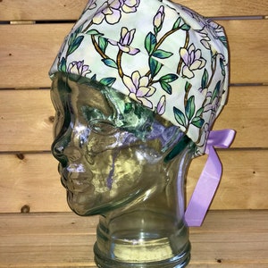 May include: A white scrub hat with a floral pattern of green leaves and purple flowers. The hat has a purple ribbon tied at the back.