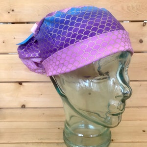 May include: A purple and blue scrub hat with a mermaid scale pattern. The hat is made of a soft, stretchy fabric and has a tie at the back.