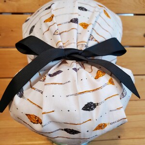 May include: White scrub hat with a black ribbon tie. The hat has a pattern of broomsticks and stars.