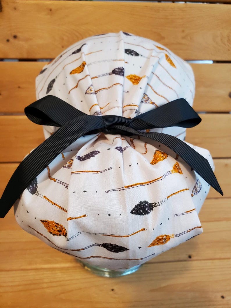 May include: White scrub hat with a black ribbon tie. The hat has a pattern of broomsticks and stars.