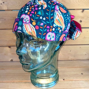 May include: A dark blue scrub hat with a colorful owl print. The hat has a pink bow tie and is made of a soft, comfortable fabric.