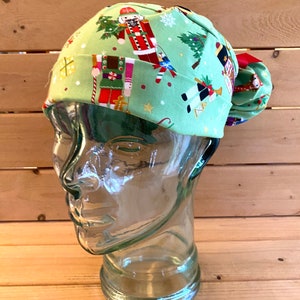 May include: A green scrub hat with a Christmas nutcracker print. The hat has a tie that is tied in a bow at the back.