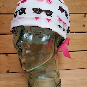 May include: White scrub hat with a pink ribbon and a pattern of black and pink sunglasses.