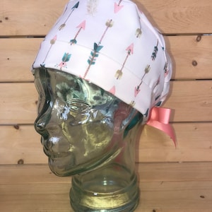 May include: A white scrub hat with a pink bow. The hat has a pattern of pink, green, and gold arrows and feathers.