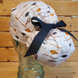 May include: A white scrub hat with a black ribbon tie. The hat has a pattern of brown and orange broomsticks on a white background.
