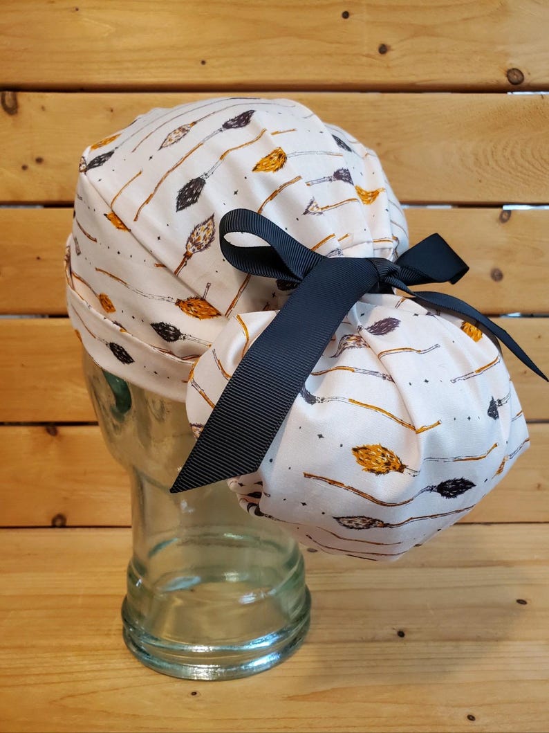 May include: A white scrub hat with a black ribbon tie. The hat has a pattern of brown and orange broomsticks on a white background.