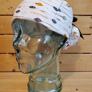 May include: White surgical scrub hat with a black tie. The hat has a pattern of gold and purple arrows on a white background.