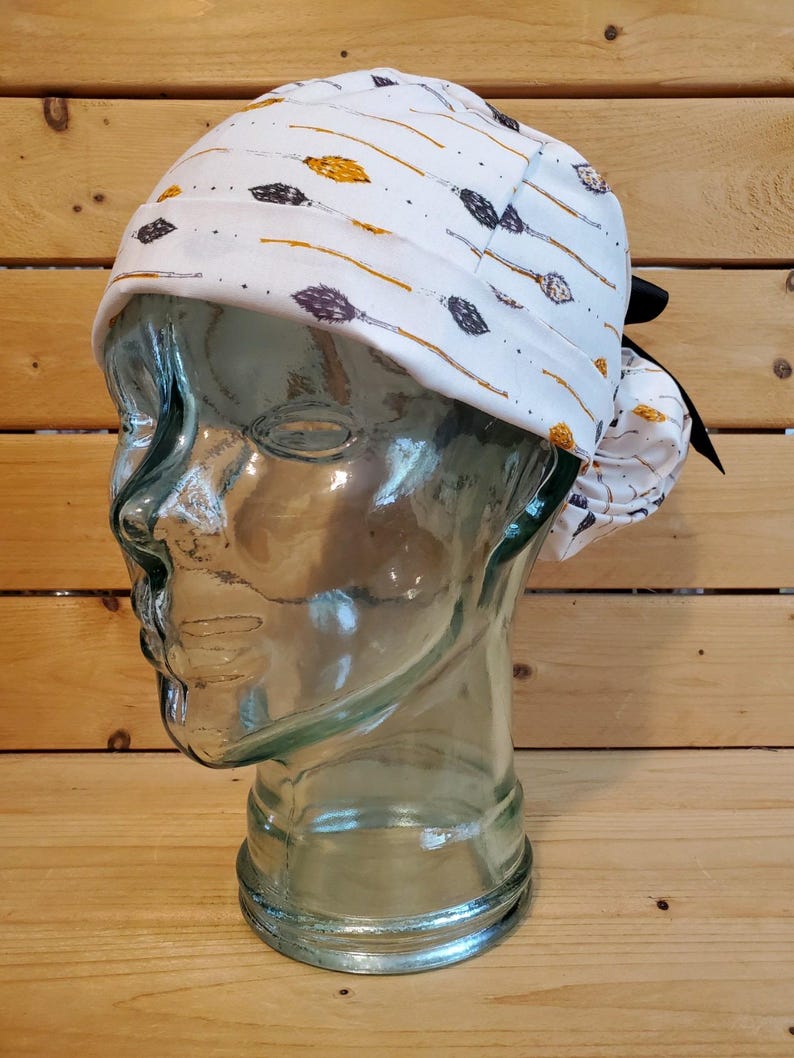 May include: White surgical scrub hat with a black tie. The hat has a pattern of gold and purple arrows on a white background.