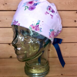 May include: White scrub cap with a floral and unicorn design. The cap features purple, pink, and blue flowers, along with unicorn horns and eyelashes. A dark blue ribbon is attached to the back of the cap.