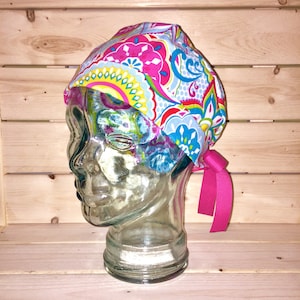 May include: A colorful patterned surgical scrub hat with a pink tie. The hat is on a glass mannequin head.