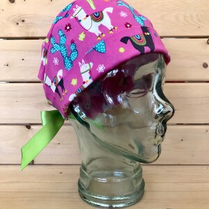 May include: A pink surgical scrub hat with a green ribbon tie. The hat has a pattern of llamas, cacti, and flowers.