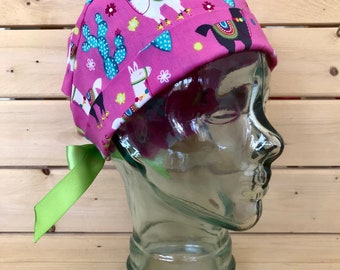 May include: A pink surgical scrub hat with a green ribbon tie. The hat has a pattern of llamas, cacti, and flowers.
