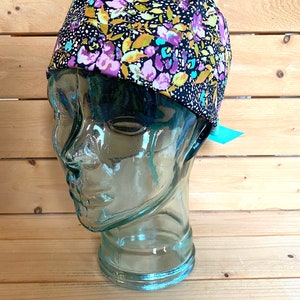 May include: A black scrub hat with a floral print in shades of purple, yellow, and teal. The hat is on a glass mannequin head.