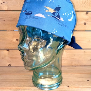 May include: A blue scrub hat with a navy blue tie. The hat has a print of ships sailing on a blue sea with white clouds and stars.