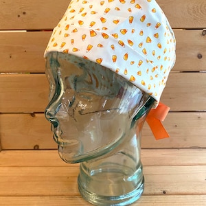 May include: White surgical scrub hat with a pattern of orange and yellow candy corn. The hat has a tie on the back.