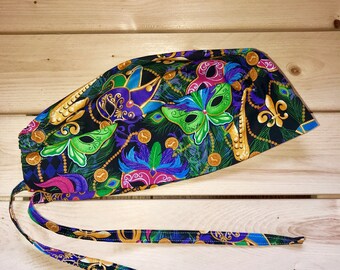 May include: A black scrub cap with a colorful Mardi Gras print featuring gold beads, green, purple, and gold masks, and fleur-de-lis. The cap has two long ties.