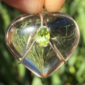 Pu&ograve; includere: Un ciondolo a forma di cuore trasparente con una pietra preziosa verde incastonata al centro. Il ciondolo &egrave; in argento e presenta un delicato design a filo.